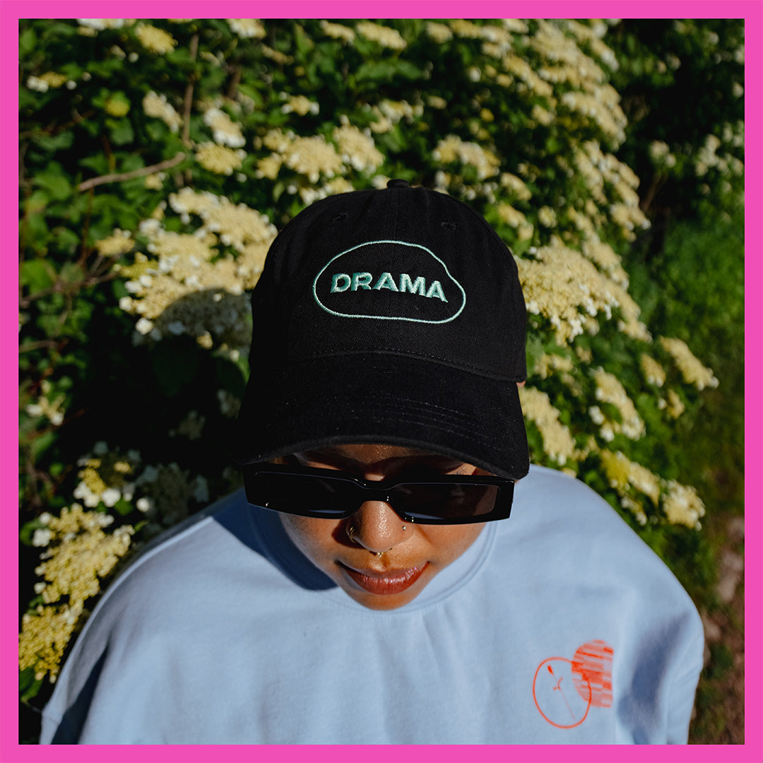 Front of black DRAMA tour cap with DRAMA embroidered in mint green, worn by Via wearing a blue crewneck and standing in greenery