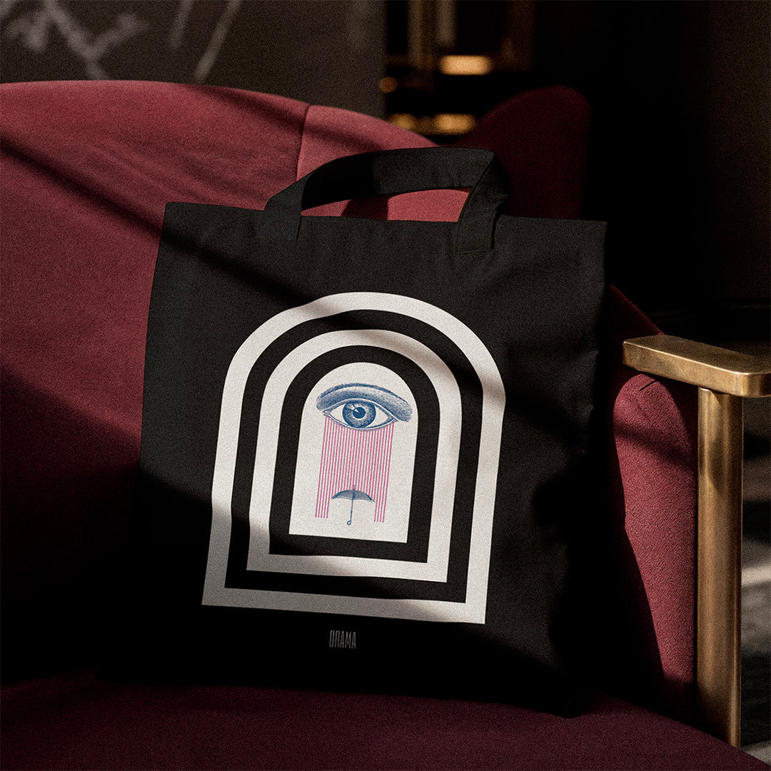 black canvas tote bag with DRAMA Dark Rain arch design printed in white, navy and pink, laying on a maroon chair