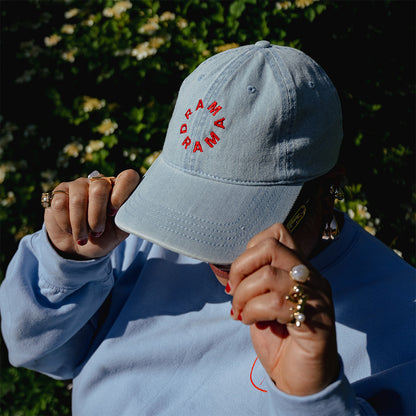 via holding the rim of the frontside of a blue denim dad cap with DRAMA embroidered in red