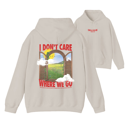 A light beige hoodie shown from both the front and back. The back features a large graphic print with the text 'I DON'T CARE WHERE WE GO' in red, surrounding an illustration of a sunlit green field seen through wooden arches with white clouds.