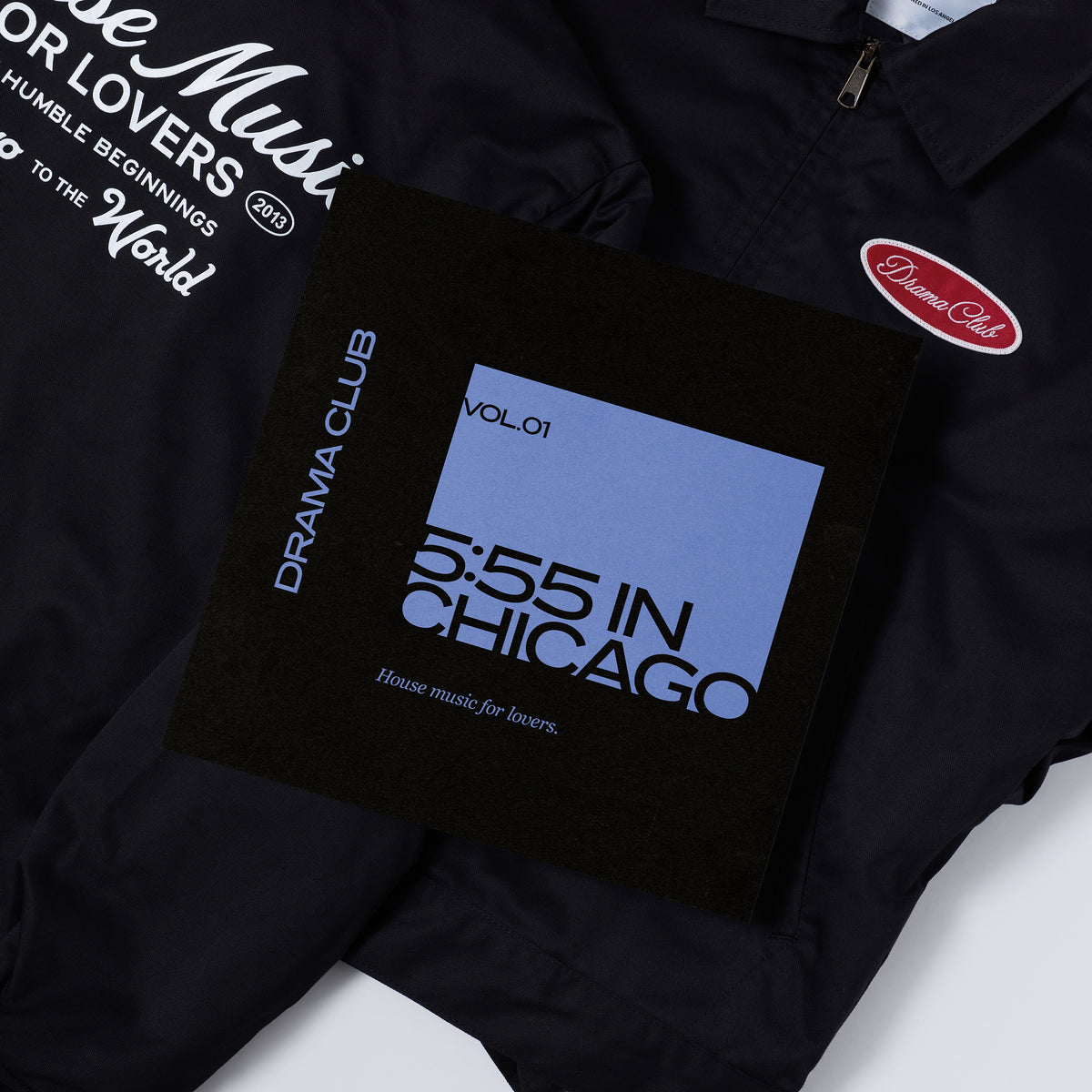 A black jacket with white printed graphics and a red oval 'Drama Club' patch, laid flat with a black vinyl cover featuring a blue panel and the text '5:55 IN CHICAGO Vol.01' and 'Drama Club'