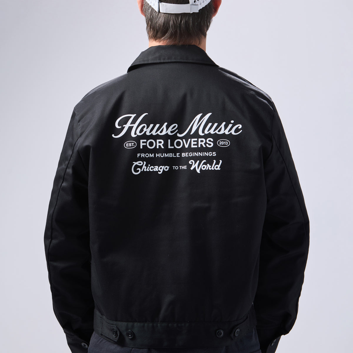 A person shown from the back wearing a black jacket with large white text on the back reading 'House Music FOR LOVERS Chicago TO THE World' and smaller text below, against a light gray studio background.