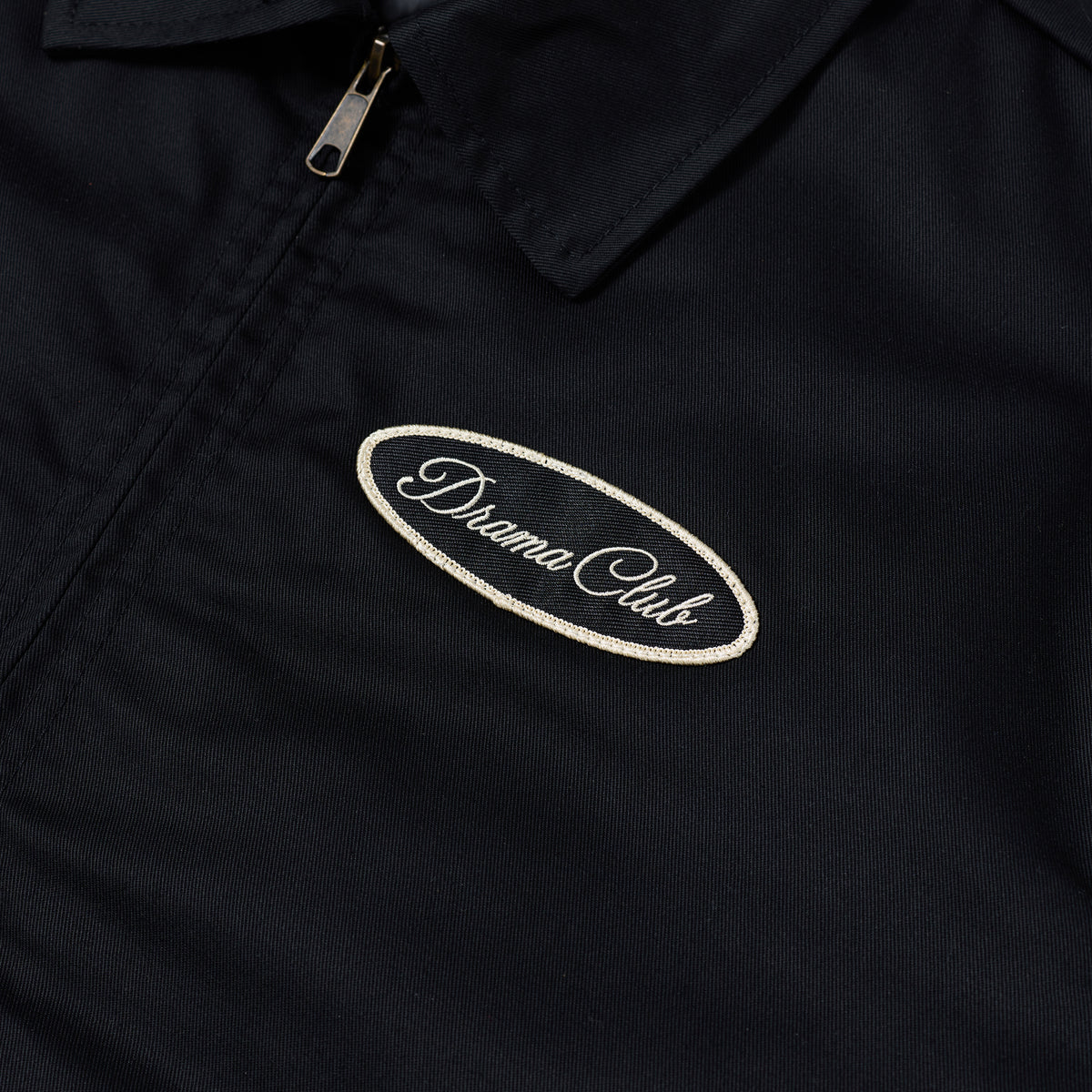 Close-up of a black zip-front jacket or overshirt with a collar and a small oval embroidered patch reading 'Drama Club' on the chest.