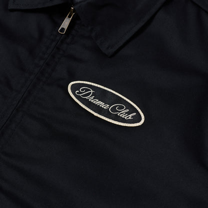 Close-up of a black zip-front jacket or overshirt with a collar and a small oval embroidered patch reading 'Drama Club' on the chest.