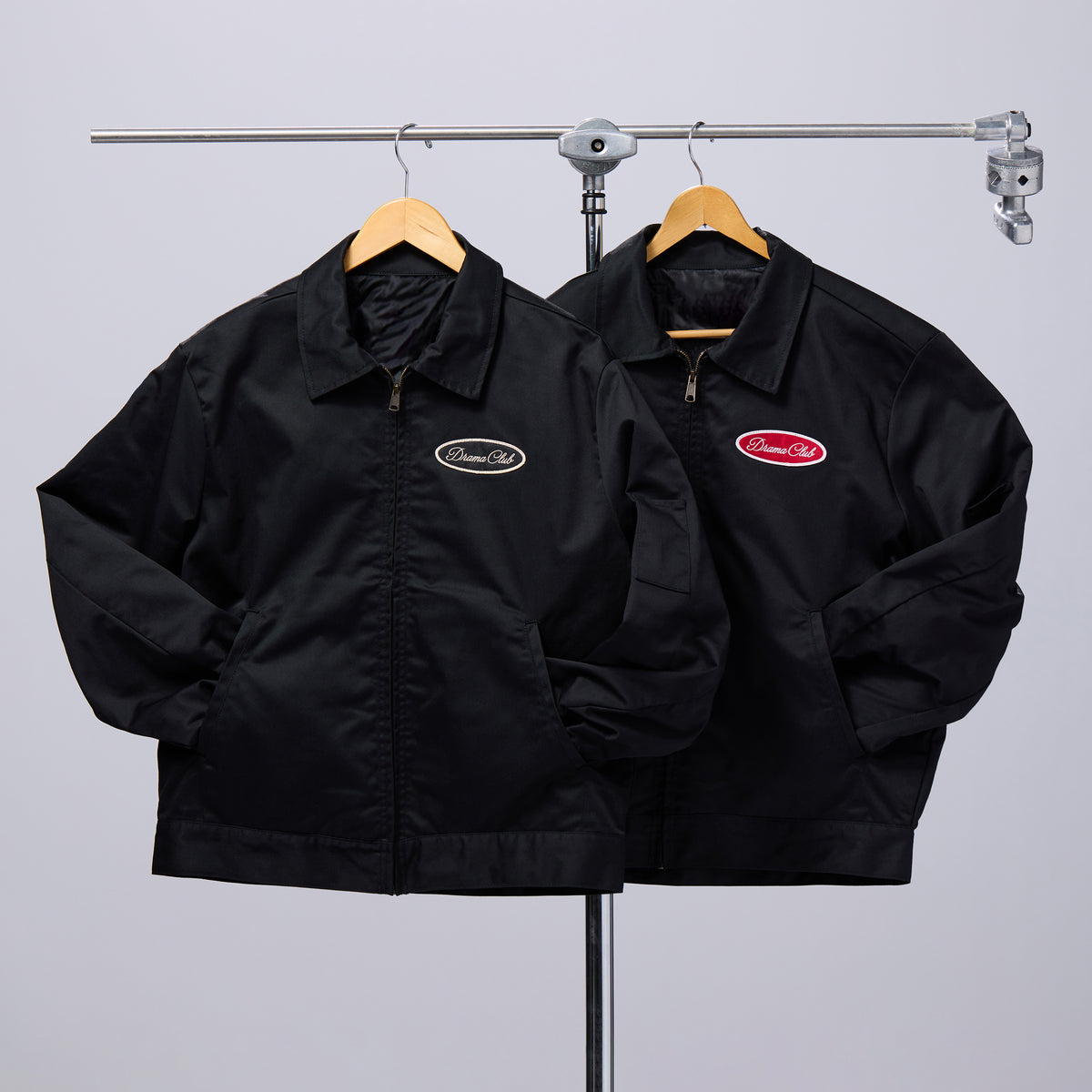 Two black zip-front jackets hanging on wooden hangers from a metal clothing rack, against a light gray studio background. The jacket on the left has a small oval logo patch on the left chest, and the jacket on the right has a larger red oval logo patch on the left chest.