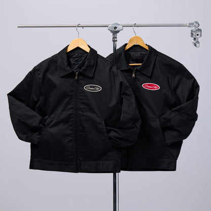 Two black zip-front jackets hanging on wooden hangers from a metal clothing rack, against a light gray studio background. The jacket on the left has a small oval logo patch on the left chest, and the jacket on the right has a larger red oval logo patch on the left chest.