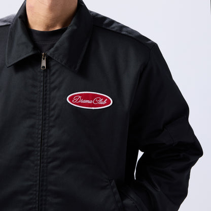 A close-up of a person wearing a black zip-up jacket with a red and white embroidered oval 'Drama Club' logo patch on the left chest, set against a plain light gray background.